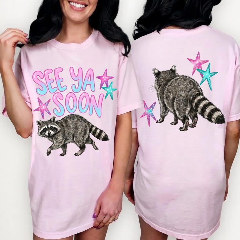 May include: Light pink t-shirt with a raccoon graphic and the text "SEE YA SOON" in a bubble font. The design includes colorful stars. The back of the shirt features a raccoon and stars.