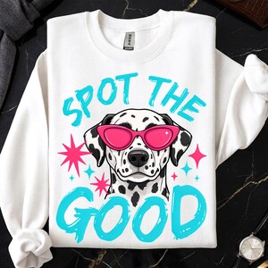 May include: A white crewneck sweatshirt featuring a graphic of a Dalmatian dog wearing bright pink cat-eye sunglasses. The text "SPOT THE GOOD" is printed in a bold, cyan brush-stroke font above and below the dog, accented by pink and cyan stars.