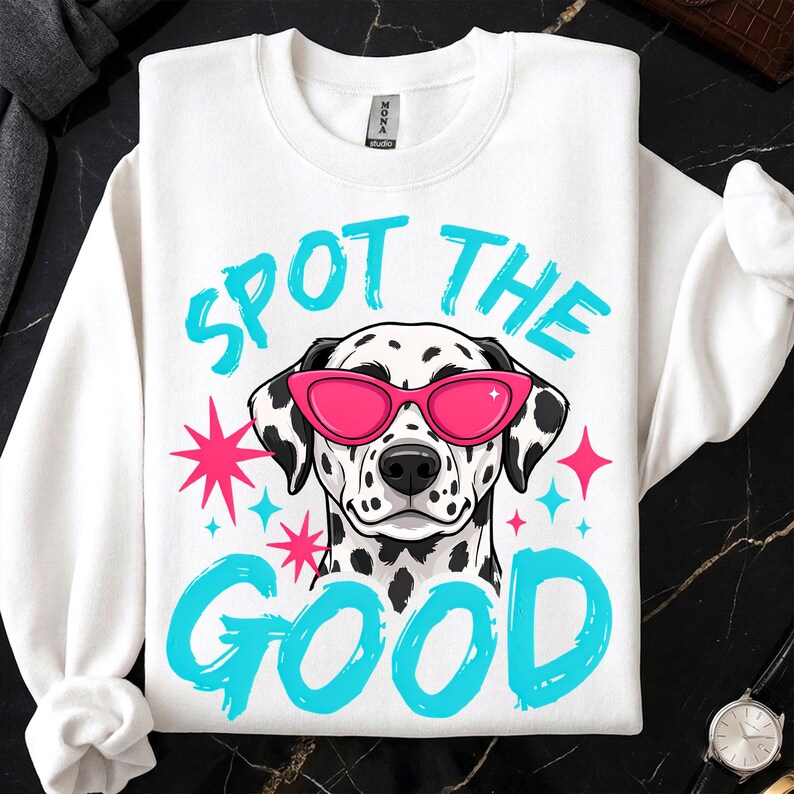 May include: A white crewneck sweatshirt featuring a graphic of a Dalmatian dog wearing bright pink cat-eye sunglasses. The text "SPOT THE GOOD" is printed in a bold, cyan brush-stroke font above and below the dog, accented by pink and cyan stars.