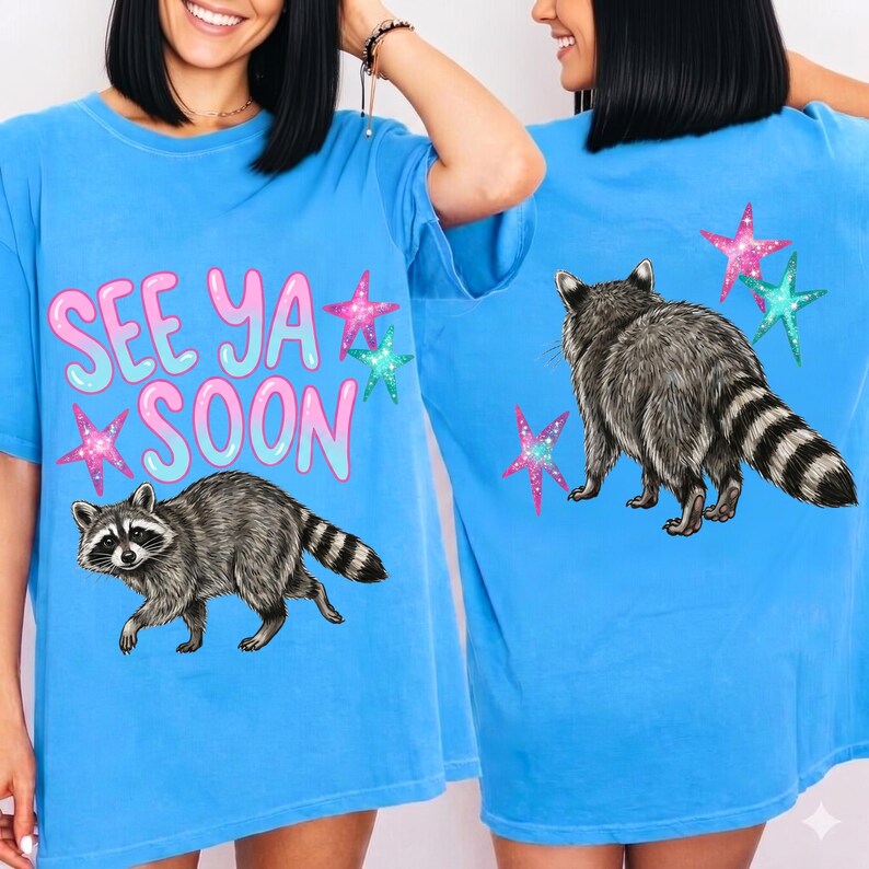 May include: Light blue t-shirt featuring a raccoon graphic and the text "SEE YA SOON" in pink and blue. The design includes star accents. The back of the shirt also features a raccoon and star graphics.