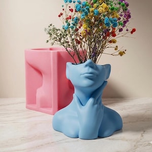 May include: A light blue ceramic face vase with a hand supporting the chin, holding colorful dried flowers. A pink rectangular vase is in the background. The vases are on a marble surface.