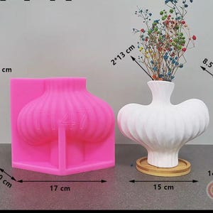 Puede incluir: Un molde de silicona rosa con forma de bulbo de ajo, de 14,5 cm de alto, junto a un jarrón de cerámica blanco con flores secas de colores. El jarrón mide 14 cm de alto y descansa sobre un posavasos de madera. El molde tiene el número "1147".