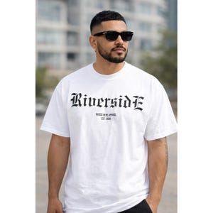 May include: White t-shirt featuring the word "Riverside" in a gothic font. Underneath, it reads "RAISED HERE APPAREL EST. 2026". The shirt has short sleeves.