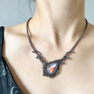 Könnte beinhalten: Eine Makramee-Halskette mit einem tropfenförmigen Labradorit-Edelstein in der Mitte, der irisierende violette und orangefarbene Reflexe aufweist. Der Stein ist in dunkelbraune, geknotete Kordel mit komplexen Webmustern und kleinen blattartigen Akzenten eingefasst.