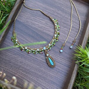 May include: A macrame necklace featuring a central teardrop-shaped blue stone pendant surrounded by intricate brown cord knotting. The necklace is adorned with small green, white, and red beads along the band, ending in two adjustable cord tails.