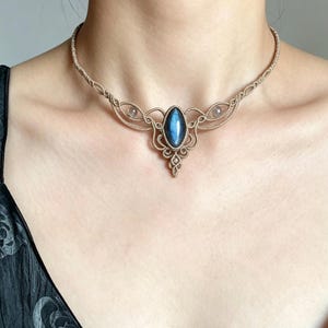 May include: A macrame choker necklace featuring a central marquise-shaped blue labradorite gemstone. The beige cord is intricately knotted in a symmetrical pattern, with two smaller round clear beads flanking the central stone on each side.