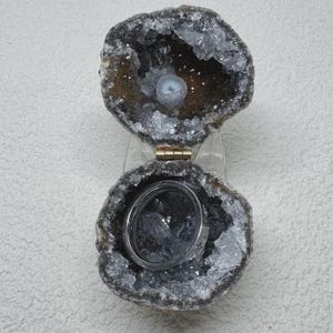 May include: A hinged geode ring box with a rough, dark brown exterior and a sparkling, crystalline interior. The geode is split into two halves connected by a gold-colored metal hinge, with a smooth, circular silver-toned ring resting inside.