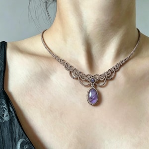 May include: A macrame necklace featuring a light brown cord woven into an intricate pattern with small metallic beads. The centerpiece is a large, oval-shaped purple gemstone, complemented by a smaller, round purple stone set above it.