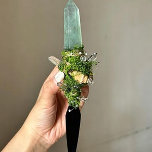 Puede incluir: Una daga decorativa con una punta de cristal verde claro translúcido, un mango envuelto en musgo verde, pequeños fragmentos de cuarzo transparente, una concha beige y un pequeño dije de estrella plateada, terminada en una hoja negra afilada.
