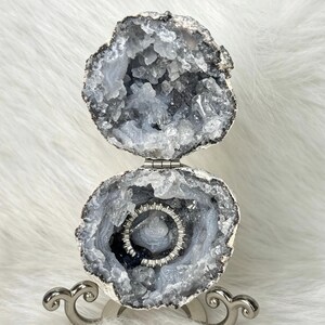 May include: A hinged geode ring box featuring a rough, grey-toned exterior and a hollow interior lined with sparkling, light blue-grey crystals. A thin, silver-toned metal band with small, clear stones is placed inside the lower geode cavity.