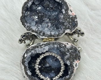 May include: A hinged geode ring box with a rough, dark gray exterior and a hollow interior lined with sparkling, light blue crystals. A thin, silver-toned eternity band with small, clear stones is resting inside the geode cavity.