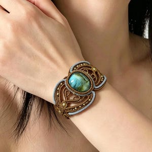 May include: A wide macrame bracelet featuring a central oval cabochon stone with blue and green hues. The bracelet is crafted from brown cord with intricate knotting patterns, accented by small metallic beads and a light blue cord border.