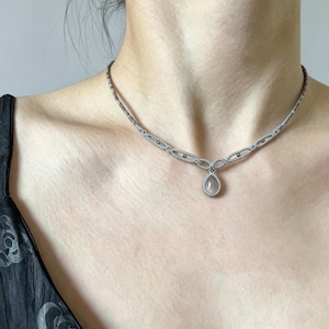 May include: A silver-toned necklace featuring a delicate chain with oval-shaped links and small embedded gold-coloured accents. The centrepiece is a teardrop-shaped pendant with a smooth, translucent grey stone set in a silver-toned frame.