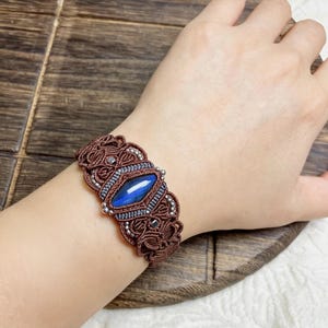 May include: A brown macrame bracelet featuring a central marquise-shaped blue gemstone. The intricate knotted cord design is accented with small metallic beads surrounding the stone, creating a detailed and textured pattern across the wrist band.