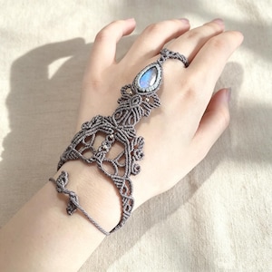 Könnte beinhalten: Ein graues Makramee-Handarmband mit einem tropfenförmigen, irisierenden Mondstein-Cabochon in der Mitte. Das aufwendige Design aus geknoteten Schnüren verbindet ein Armband mit einem Fingerring zu einem dekorativen Schmuckstück für die Hand.
