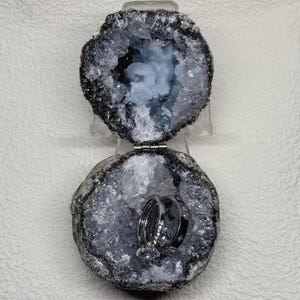 Puede incluir: Un joyero de geoda con bisagra, exterior gris oscuro rugoso y cavidad interior cristalina brillante. La geoda está abierta, revelando dos anillos de metal plateado que descansan dentro del centro hueco que refleja la luz.