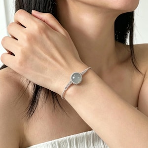 May include: A bracelet featuring a central round, pale green, translucent cabochon stone set in a silver-toned metal bezel with a beaded edge. The band consists of a delicate, woven silver-toned chain that connects to the central stone setting.