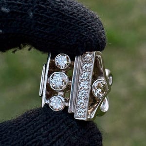 May include: A silver ring with multiple clear gemstones. The ring features a row of small stones and several larger, round stones. The ring is held by a black glove against a blurred green background.