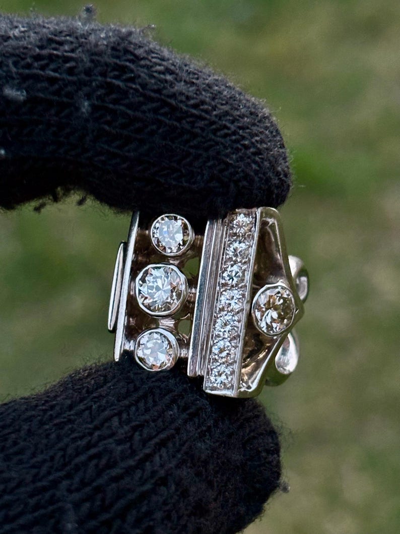May include: A silver ring with multiple clear gemstones. The ring features a row of small stones and several larger, round stones. The ring is held by a black glove against a blurred green background.