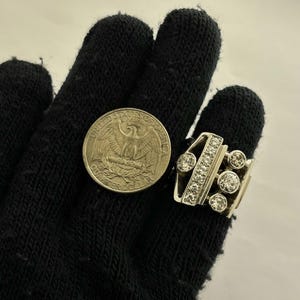 May include: A silver-toned ring with multiple clear gemstones and a quarter coin. The ring features a rectangular design with a row of small stones and several larger, round stones. The quarter coin is visible next to the ring.