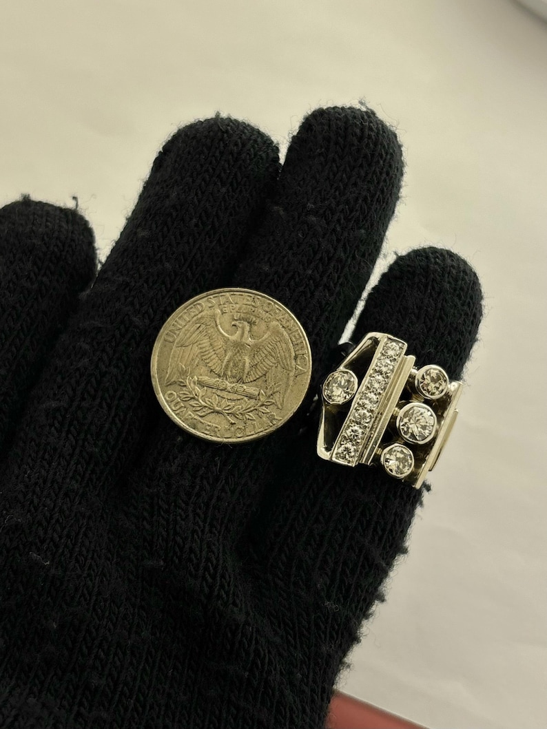 May include: A silver-toned ring with multiple clear gemstones and a quarter coin. The ring features a rectangular design with a row of small stones and several larger, round stones. The quarter coin is visible next to the ring.
