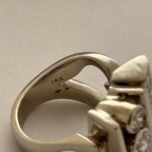 May include: A close-up of a gold-colored ring with a unique design. The ring features three clear, round gemstones and the inscription "14K". The ring has a distinctive shape with a curved band and an ornate setting for the gemstones.