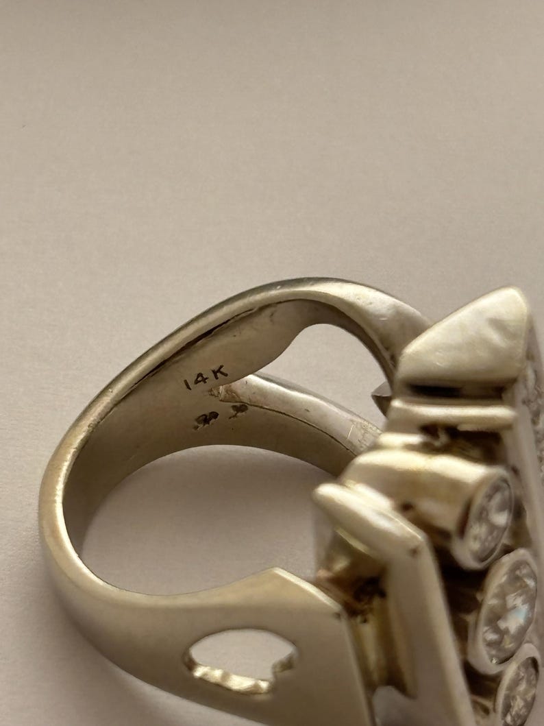 May include: A close-up of a gold-colored ring with a unique design. The ring features three clear, round gemstones and the inscription "14K". The ring has a distinctive shape with a curved band and an ornate setting for the gemstones.