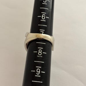 May include: A silver ring is placed on a black ring sizer. The ring is a size 7. The ring is a simple band design. The ring sizer has measurements from 5 to 10.