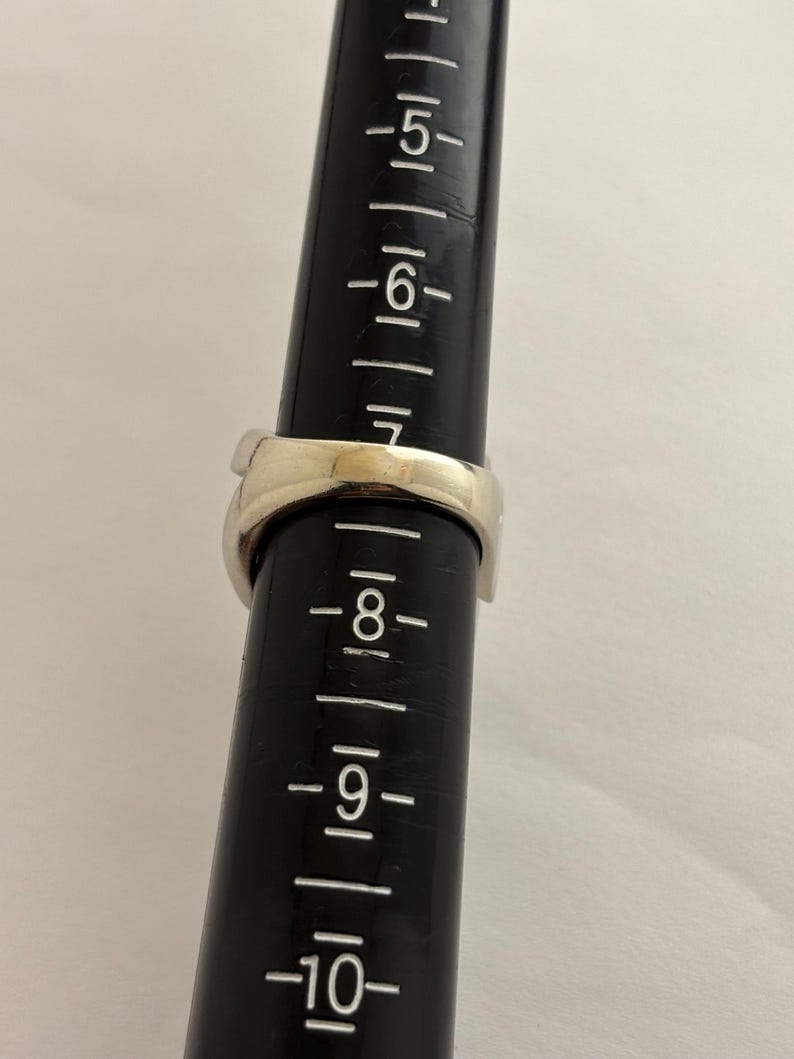 May include: A silver ring is placed on a black ring sizer. The ring is a size 7. The ring is a simple band design. The ring sizer has measurements from 5 to 10.