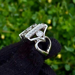May include: A silver ring with a unique design, featuring multiple small diamonds and a larger central stone. The ring has a heart-shaped detail on the band. The ring is displayed against a green background.