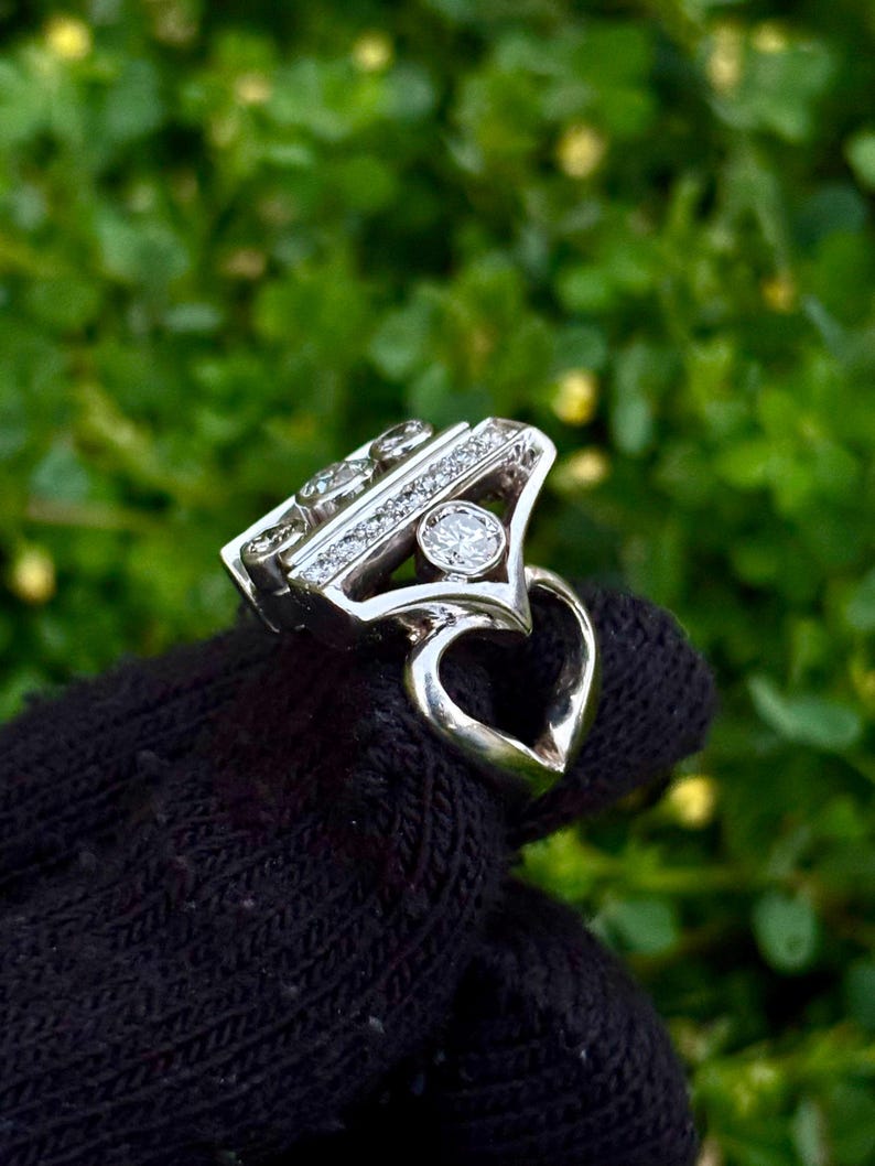 May include: A silver ring with a unique design, featuring multiple small diamonds and a larger central stone. The ring has a heart-shaped detail on the band. The ring is displayed against a green background.