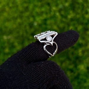 May include: A silver ring with a heart-shaped base and a rectangular top adorned with multiple clear gemstones. The ring is held up against a green background, showcasing its intricate design and sparkling details.