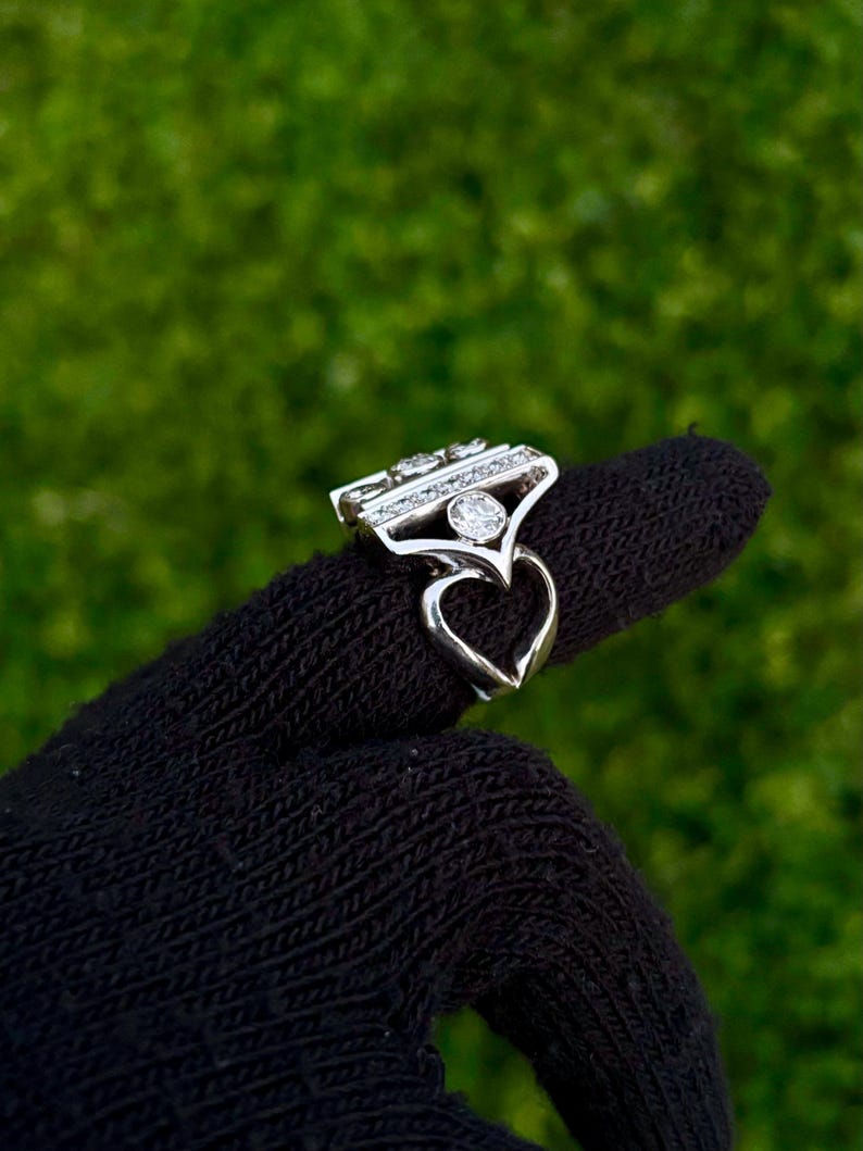 May include: A silver ring with a heart-shaped base and a rectangular top adorned with multiple clear gemstones. The ring is held up against a green background, showcasing its intricate design and sparkling details.
