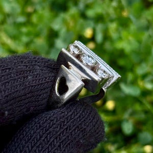 May include: A silver ring with a rectangular top featuring three round, clear gemstones and smaller stones. The ring has a heart-shaped cutout on the side. The ring is held against a dark background of a knitted fabric, with a blurred green background.