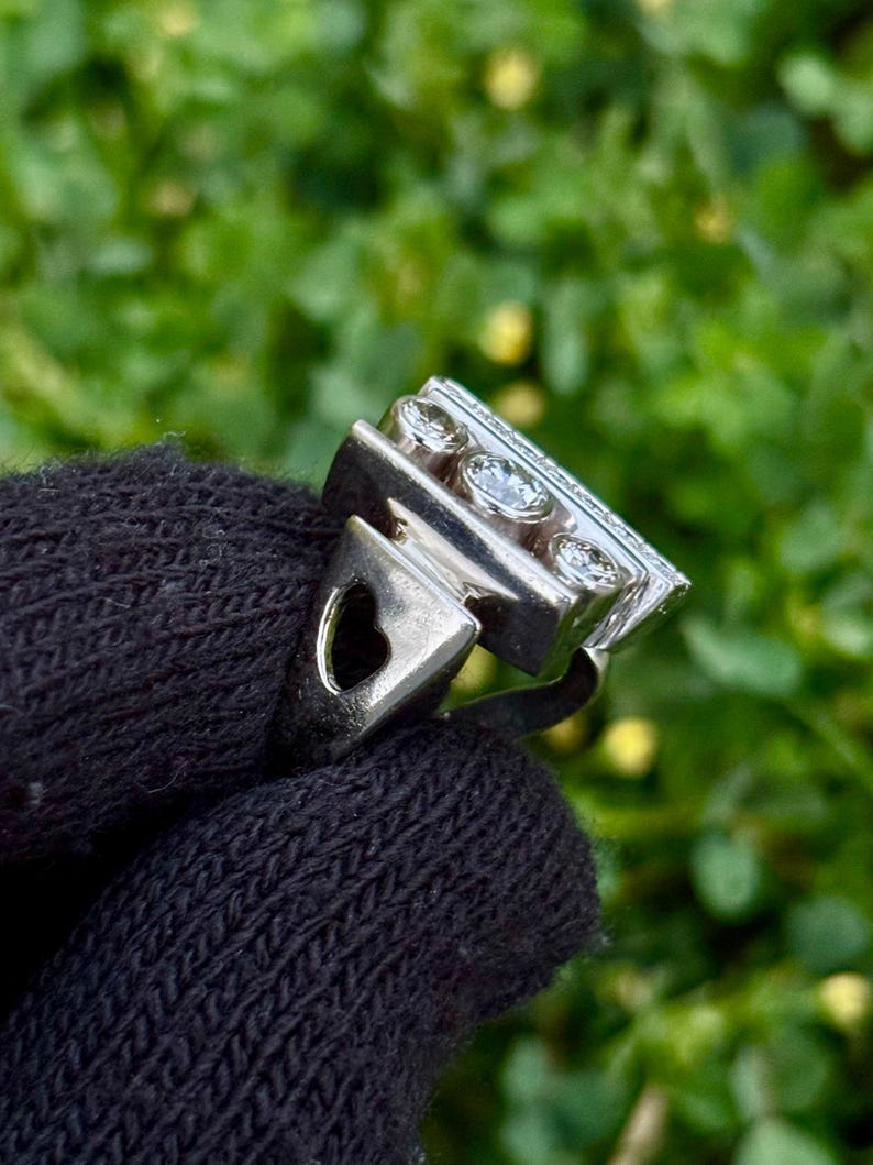 May include: A silver ring with a rectangular top featuring three round, clear gemstones and smaller stones. The ring has a heart-shaped cutout on the side. The ring is held against a dark background of a knitted fabric, with a blurred green background.