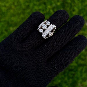May include: A silver ring with multiple clear gemstones. The ring features a rectangular design with a row of small stones and several larger, round stones. The ring is displayed on a black surface against a green background.