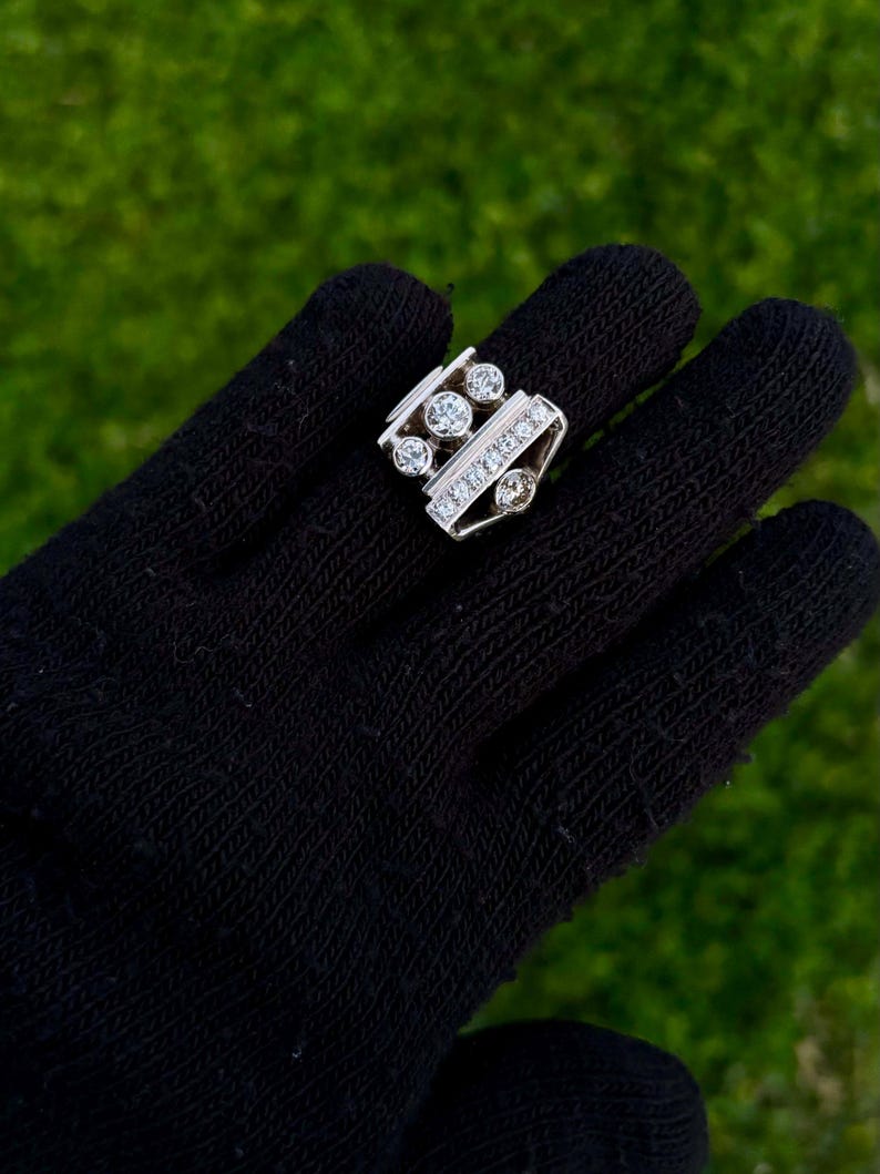 May include: A silver ring with multiple clear gemstones. The ring features a rectangular design with a row of small stones and several larger, round stones. The ring is displayed on a black surface against a green background.