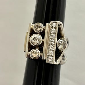 May include: A silver ring with a geometric design. The ring features several round, clear gemstones and a row of smaller stones. The ring is displayed on a black stand against a white background.