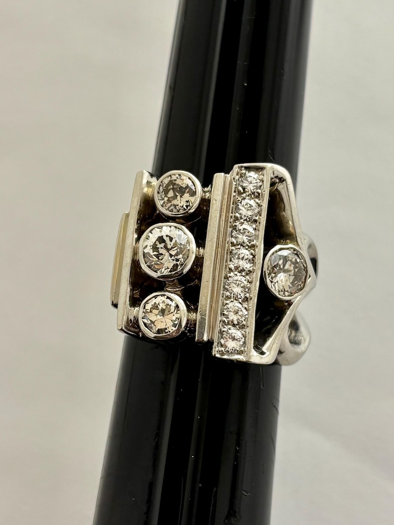 May include: A silver ring with a geometric design. The ring features several round, clear gemstones and a row of smaller stones. The ring is displayed on a black stand against a white background.