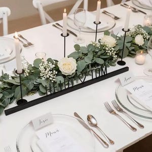 Könnte beinhalten: Eine Hochzeitstafel mit weißer Tischdecke, Blumenschmuck, Kerzen und Gedecken. Ein schwarzes Metallschild schreibt "HEIDI <3 TOM". Klare Glasplatten, Besteck und Namenskarten vervollständigen die elegante Dekoration.