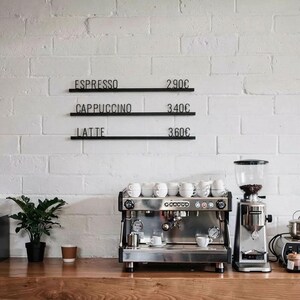 Könnte beinhalten: Ein Café-Interieur mit einer Espressomaschine aus Edelstahl, einer Kaffeemühle und weißen Kaffeetassen. Schwarze Schilder an der weißen Backsteinmauer zeigen Kaffeeoptionen: Espresso, Cappuccino und Latte.