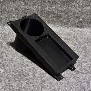 May include: Black angled console with a circular cup holder and a rectangular tray. The console has mounting tabs and is made of a textured material. The design is simple and functional.