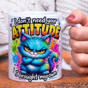 May include: White ceramic mug featuring a grumpy blue cat illustration with rainbow accents and the text "I don't need your ATTITUDE, I brought my own." The mug has a white handle and is held by a person.