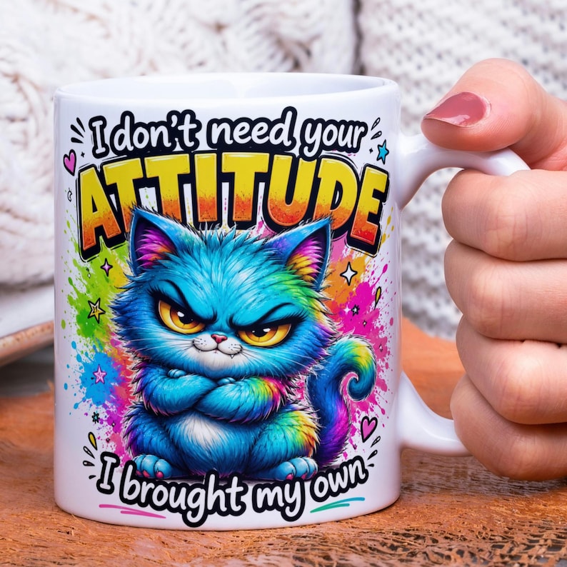 May include: White ceramic mug featuring a grumpy blue cat illustration with rainbow accents and the text "I don't need your ATTITUDE, I brought my own." The mug has a white handle and is held by a person.