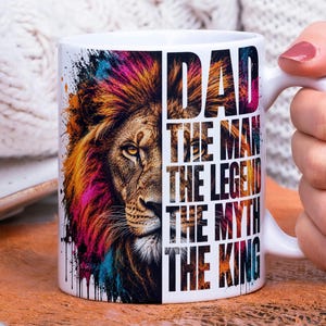 May include: White ceramic mug featuring a colorful lion's face with text that reads "DAD THE MAN THE LEGEND THE MYTH THE KING." The lion's mane has splashes of pink, orange, and blue. The mug has a white handle.