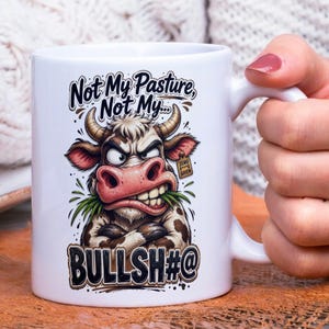 Op de afbeelding: Witte keramische mok met een humoristische afbeelding van een boze koe. De koe heeft bruine en witte vlekken, een roze neus en houdt gras in haar mond. De tekst op de mok luidt: "Not My Pasture, Not My... BULLSH#@".