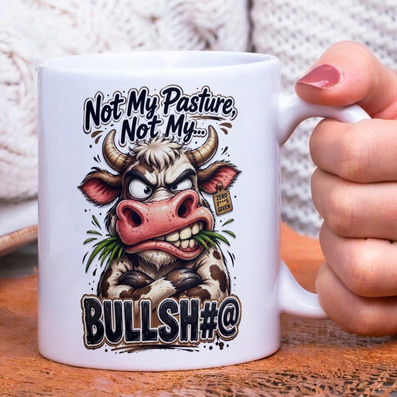 Op de afbeelding: Witte keramische mok met een humoristische afbeelding van een boze koe. De koe heeft bruine en witte vlekken, een roze neus en houdt gras in haar mond. De tekst op de mok luidt: "Not My Pasture, Not My... BULLSH#@".