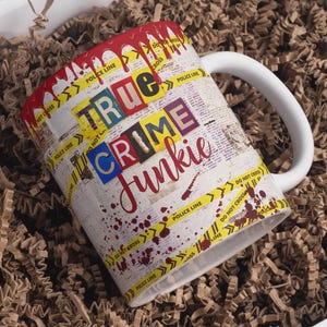 以下が含まれることがあります： 「TRUE CRIME Junkie」の文字がコラージュ風にデザインされた白いセラミックマグカップ。新聞の印刷背景、黄色の警察テープ、赤い血しぶきのディテールが特徴です。ハンドルは白です。
