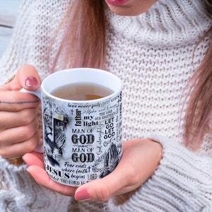 May include: White ceramic mug with a religious theme. The mug features the words "MAN OF GOD" and "JESUS" in a black and white design. The mug is filled with a light brown liquid.