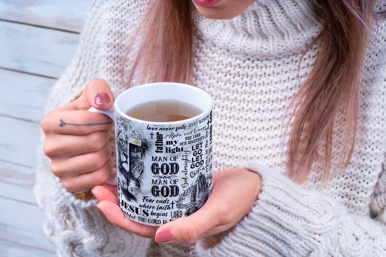 May include: White ceramic mug with a religious theme. The mug features the words "MAN OF GOD" and "JESUS" in a black and white design. The mug is filled with a light brown liquid.